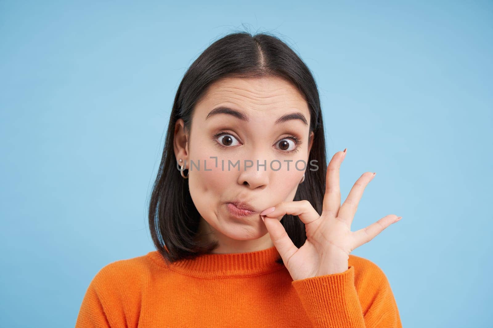 Gossips. Close up of cute asian girl zips mouth, locks lips on seal, promise to keep secret, stands over blue background.
