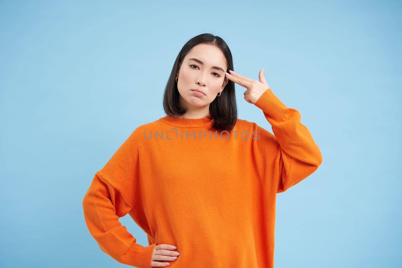 Portrait of asian woman annoyed by something, holds two fingers near head temple, bang bang gesture, stands over blue background by Benzoix