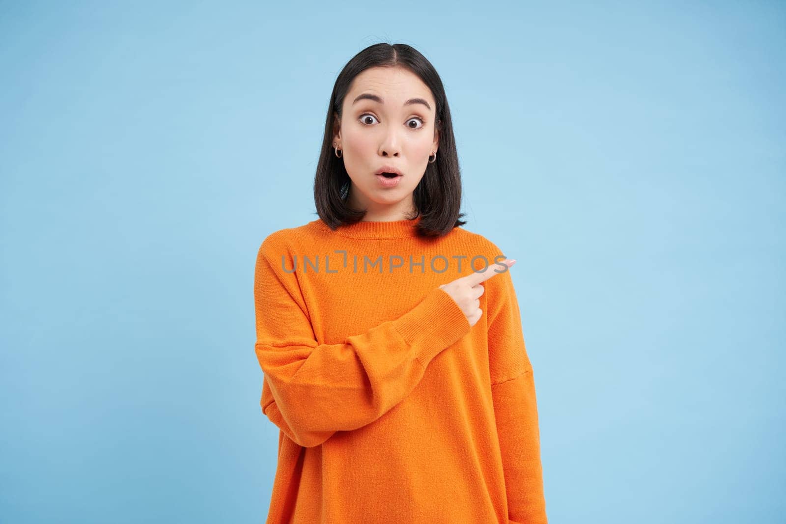 Surprised korean woman, pointing right, showing promo banner with amazed face expression, stands over blue background. Copy space