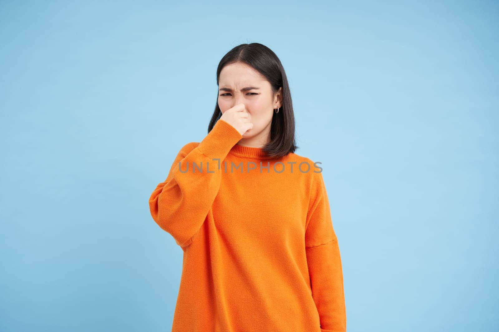 Something stinks. Young asian woman shuts her nose with fingers from bad smell, wears orange sweatshirt, stands over blue background.