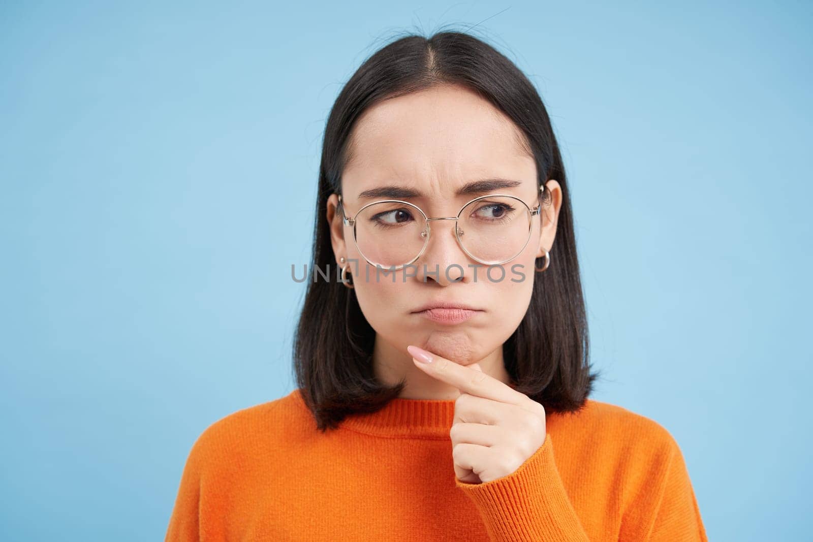 Portrait of thoughtful asian woman in glasses, girl in prescribed eyewear thinking, frowns while looks suspicious, stands over blue background.