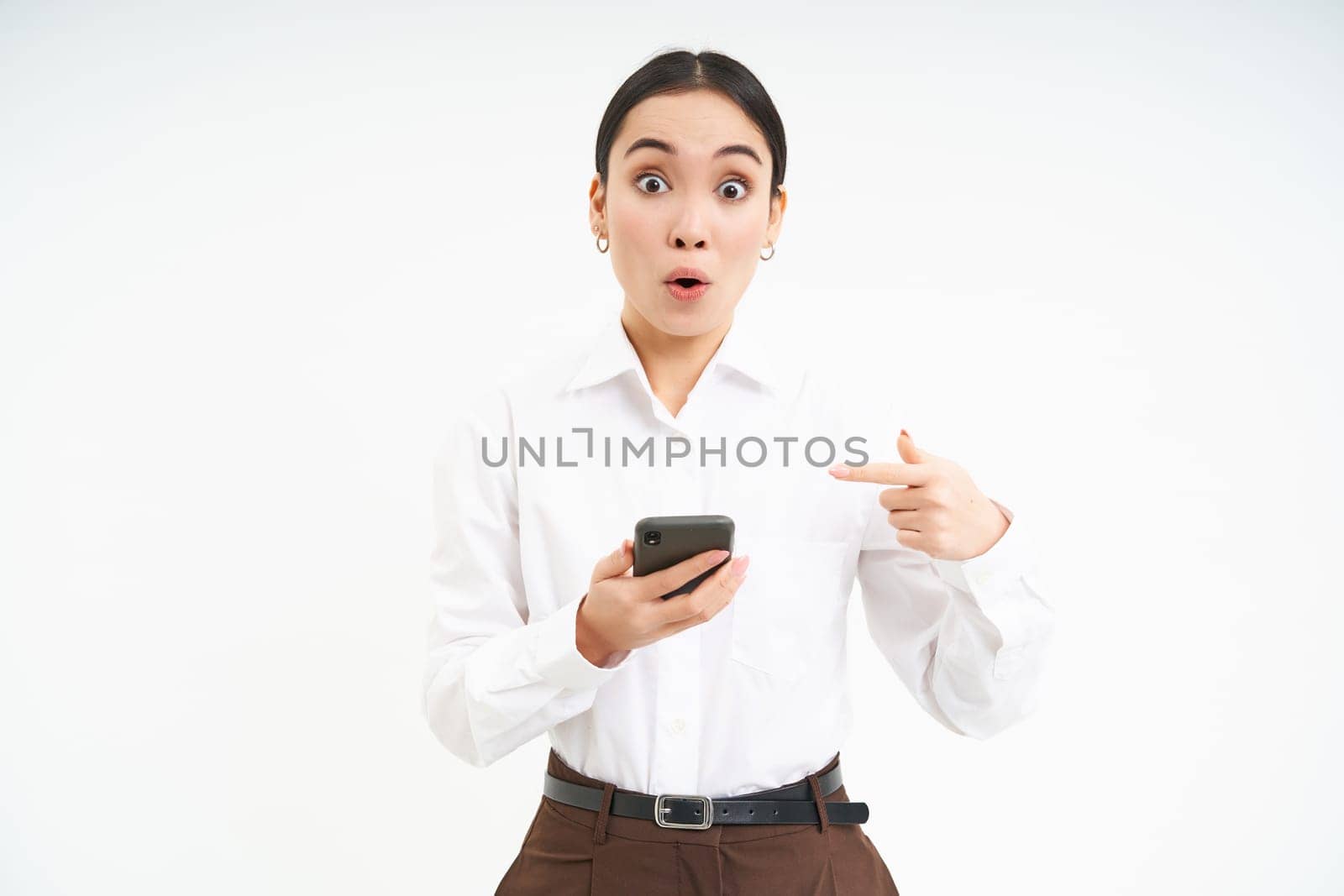 Business concept. Impressed asian corporate woman, pointing at mobile phone with surprised face, amazed by smartphone app, standing over white background by Benzoix
