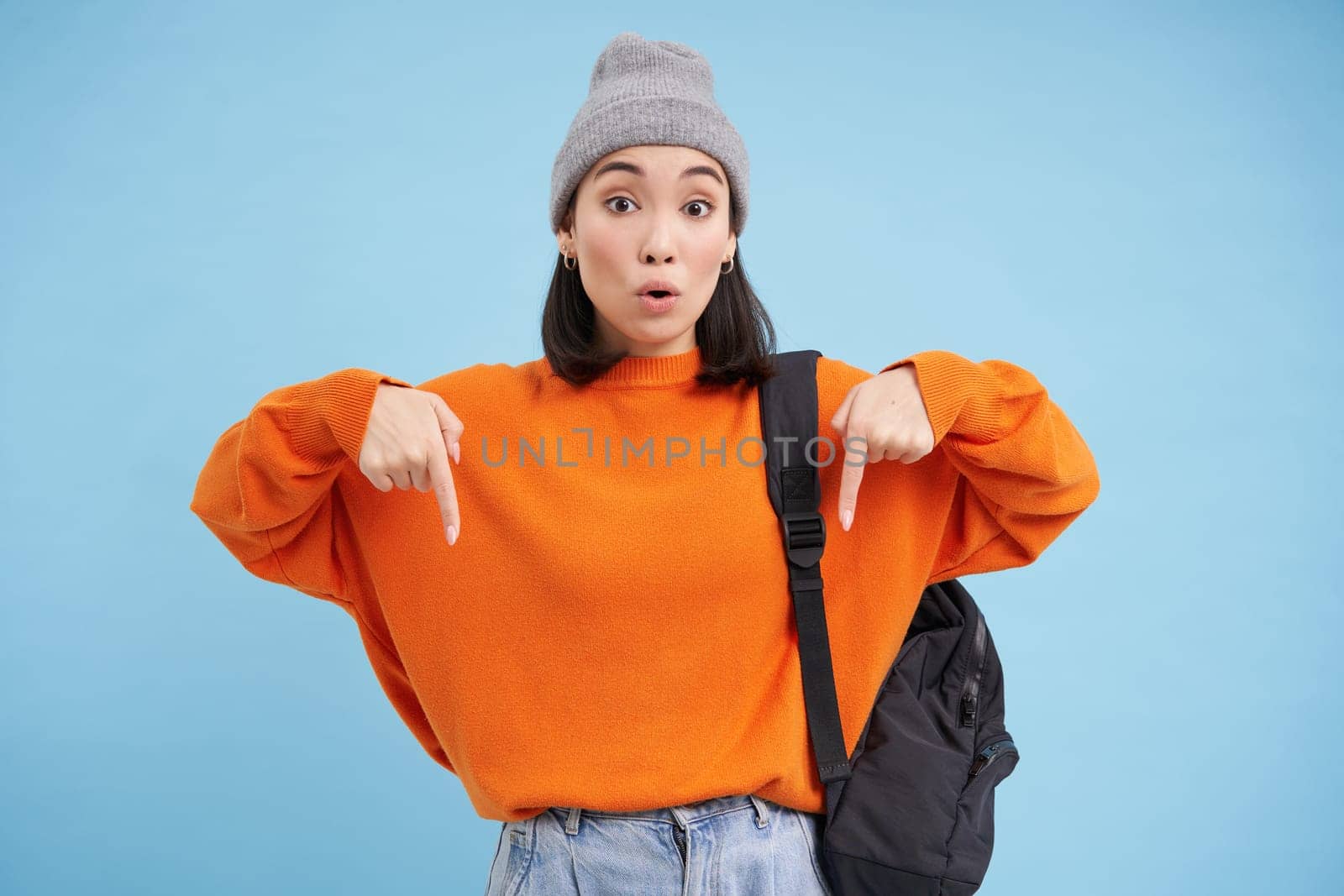 Asian woman in warm hat, wears street outfit and backpack, points fingers down, shows advertisement below, standing over blue background.