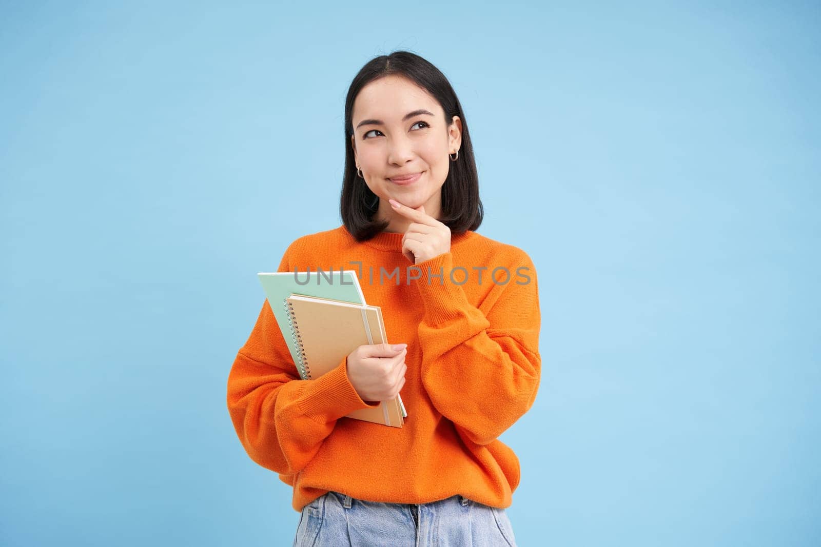 Thinking girl, student with notebooks, stands over blue background thoughtful, making decision.