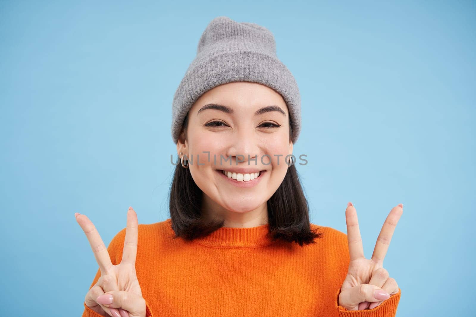 Close up portrait of positive and cute asian girl in beanie, smiles and shows peace, kawaii sign blue background by Benzoix