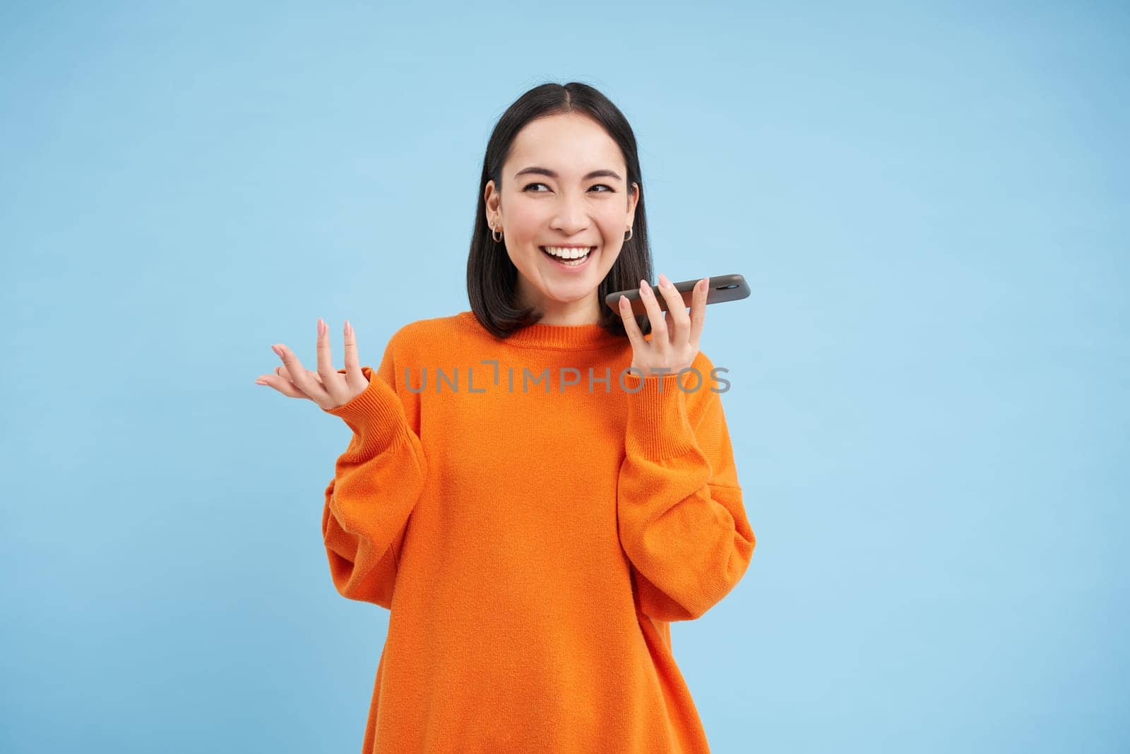 Happy asian woman records voice message, repeats words in language learning mobile app, talks in smartphone speakerphone, blue background by Benzoix