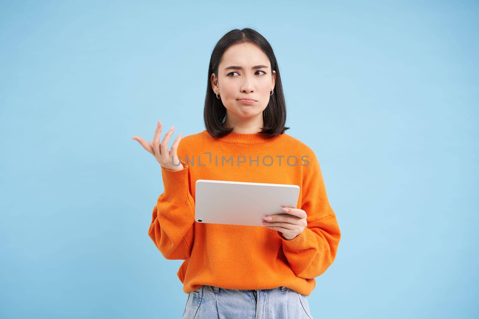 Woman with digital tablet, looks confused, cant understand smth on her gadget, stands puzzled against blue background by Benzoix