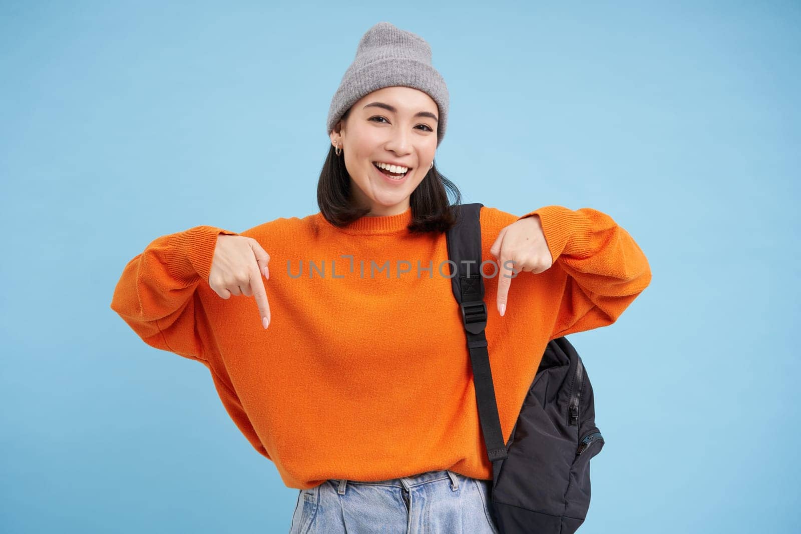 Asian woman in warm hat, wears street outfit and backpack, points fingers down, shows advertisement below, standing over blue background.