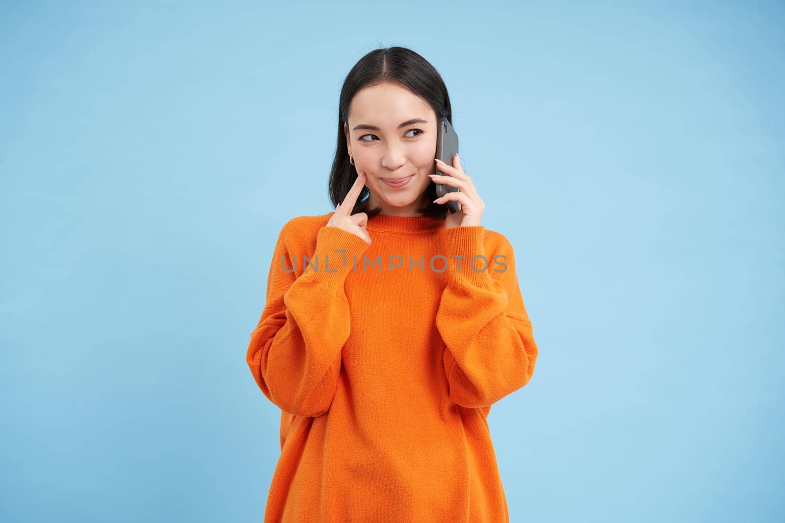 Young woman with thoughtful look, answers phone call, talks on mobile and smiling, thinking with pleased face, standing over blue background.