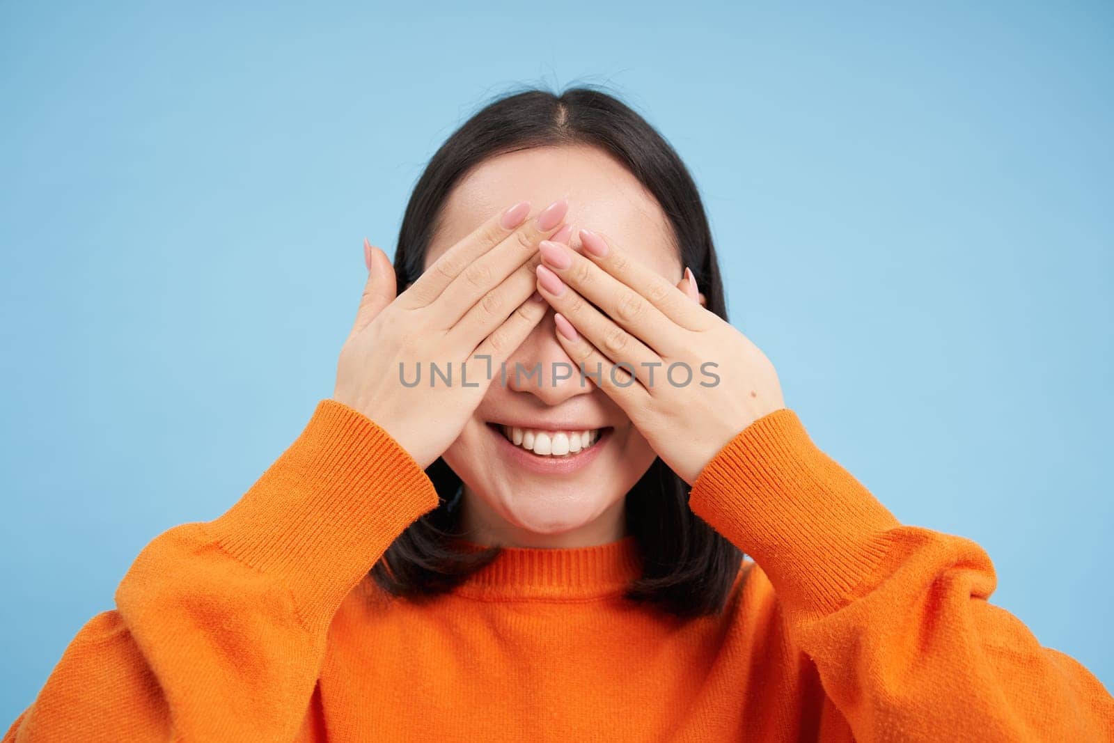 Close up of asian woman shuts her eyes and smiles blindfolded, waits for surprise, stands over blue background by Benzoix