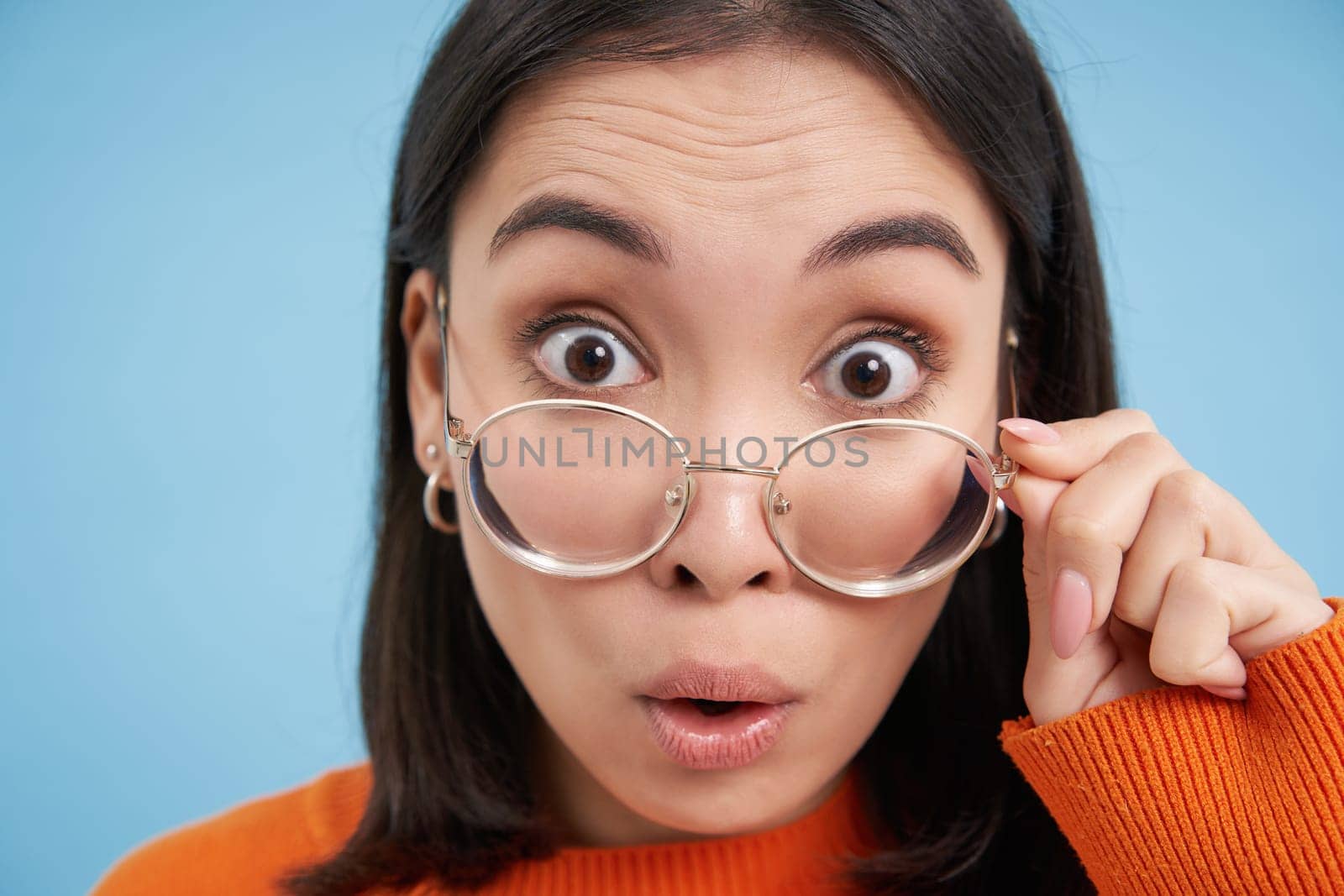 Portrait of amazed japanese woman in glasses, reads smth interesting, wears prescribed eyewear, blue background.