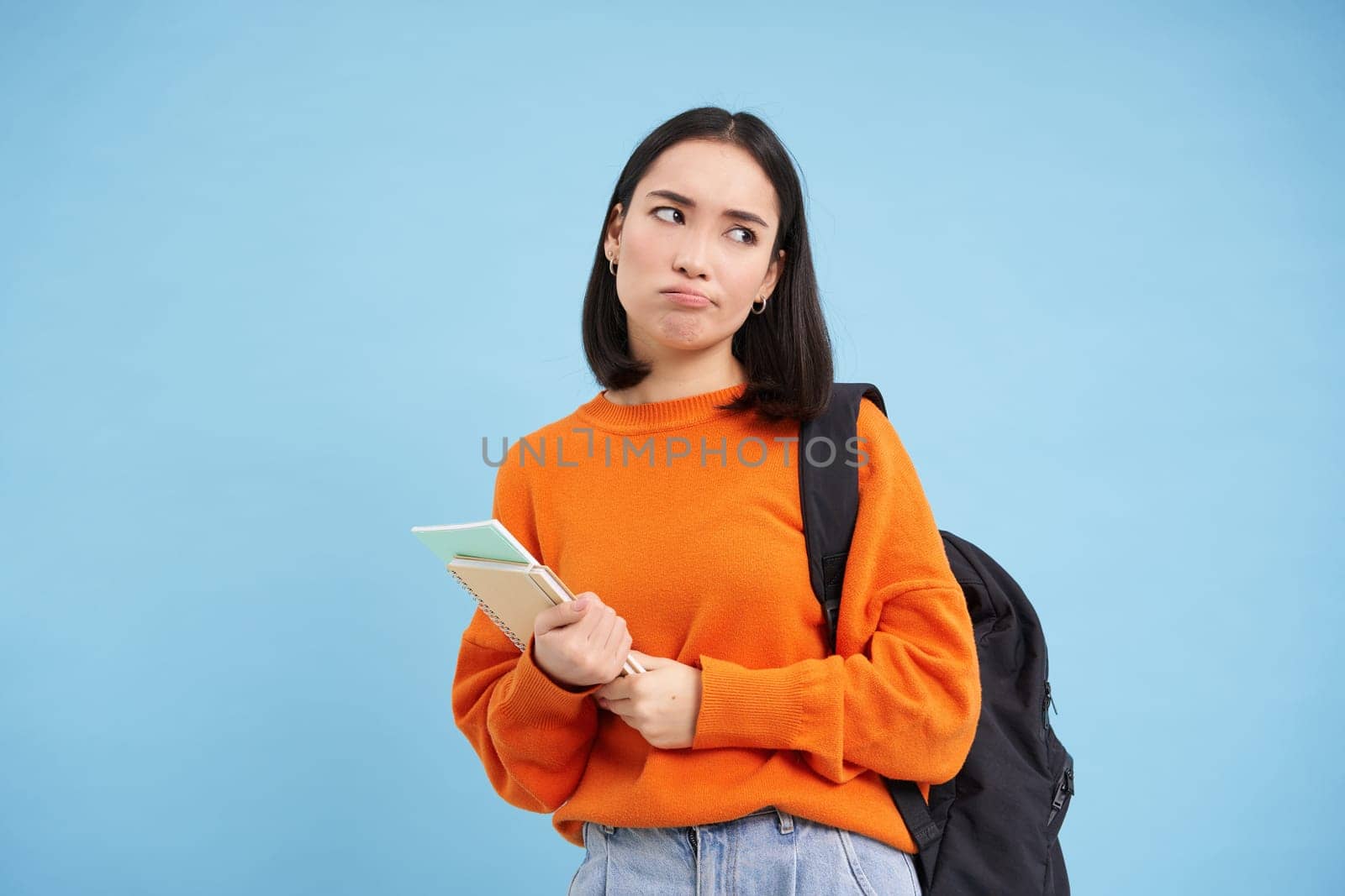 Bored college student, girl with backpack and notebooks, sulks and rolls eyes, stands upset against blue background.