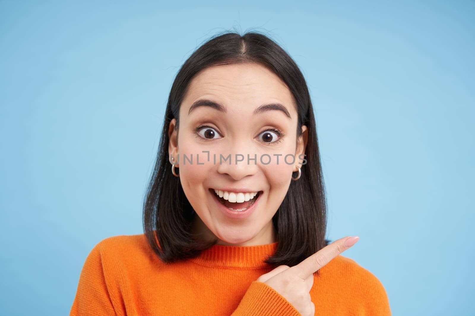 Close up portrait of smiling asian woman, pointing finger right, showing advertisement, copy space banner, standing over blue background by Benzoix