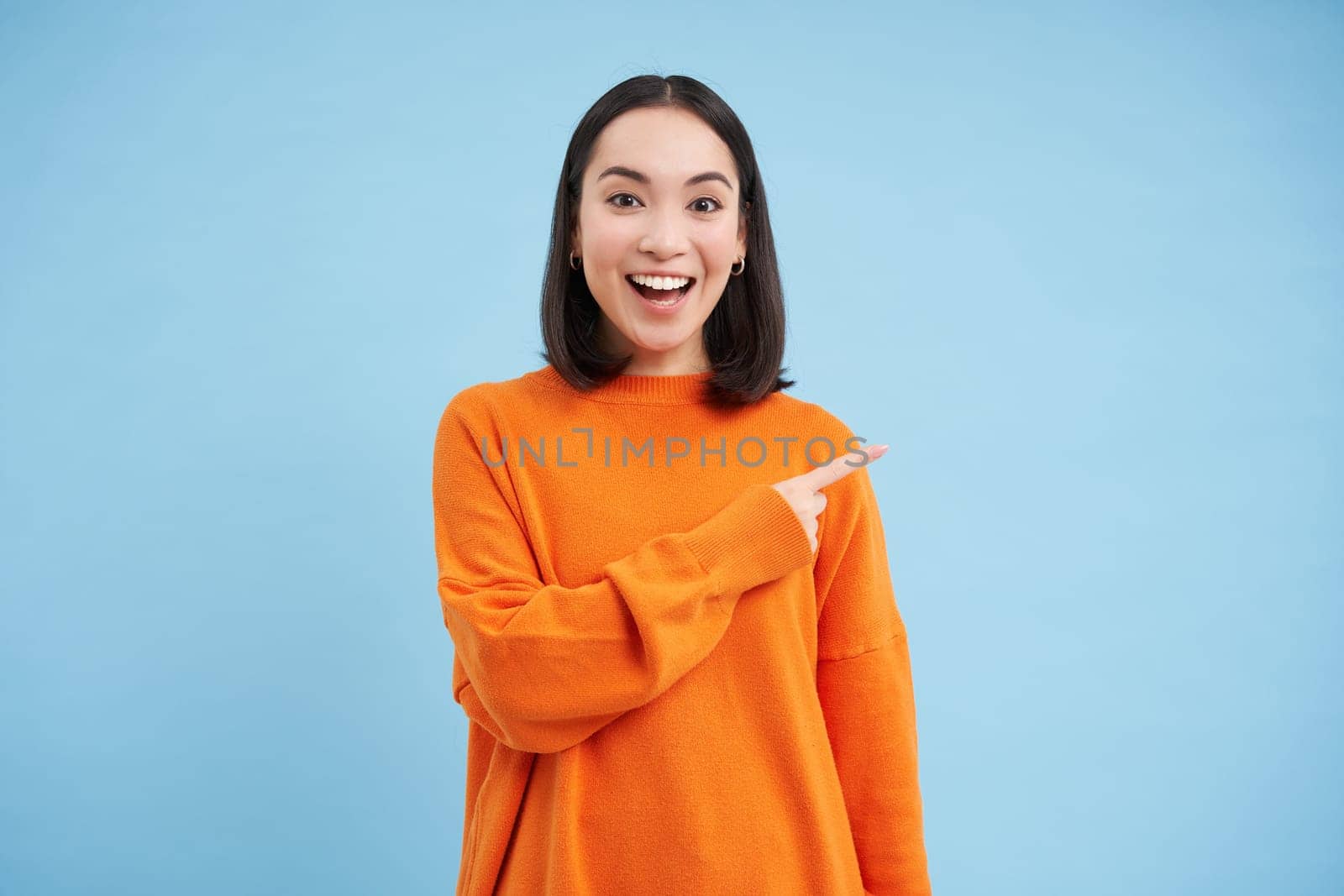 Happy smiling asian woman, pointing finger to the right, shows advertisement, blank copy space on blue background.
