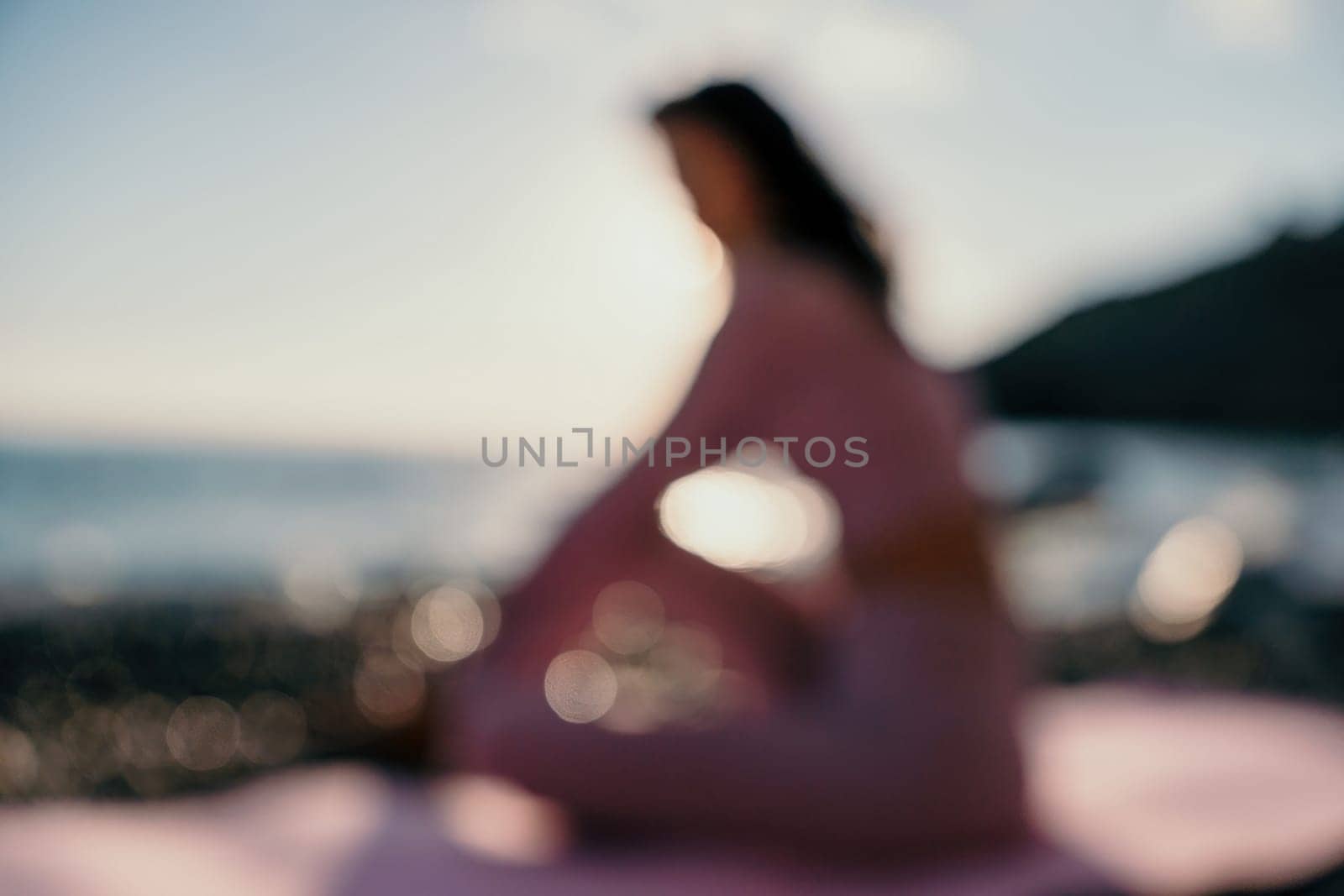 Abstract defocused silhoute of middle aged well looking woman with black hair doing Pilates with the ring on the yoga mat near the sea on the pebble beach. Female fitness yoga concept. by panophotograph