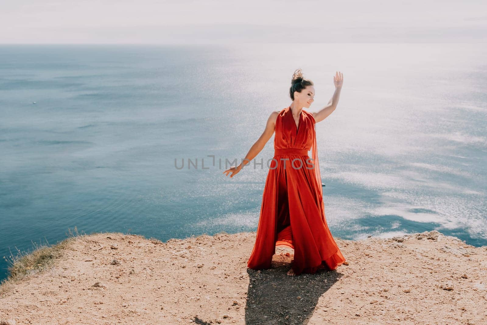 Woman in red dress on sea. Side view a Young beautiful sensual woman in a red long dress posing on a rock high above the sea on sunset. Girl on the nature on blue sky background. Fashion photo. by panophotograph