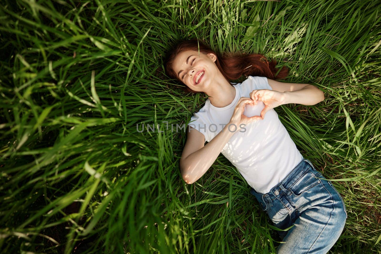 happy woman folded her arms on her chest in the shape of a heart lying in the grass by Vichizh