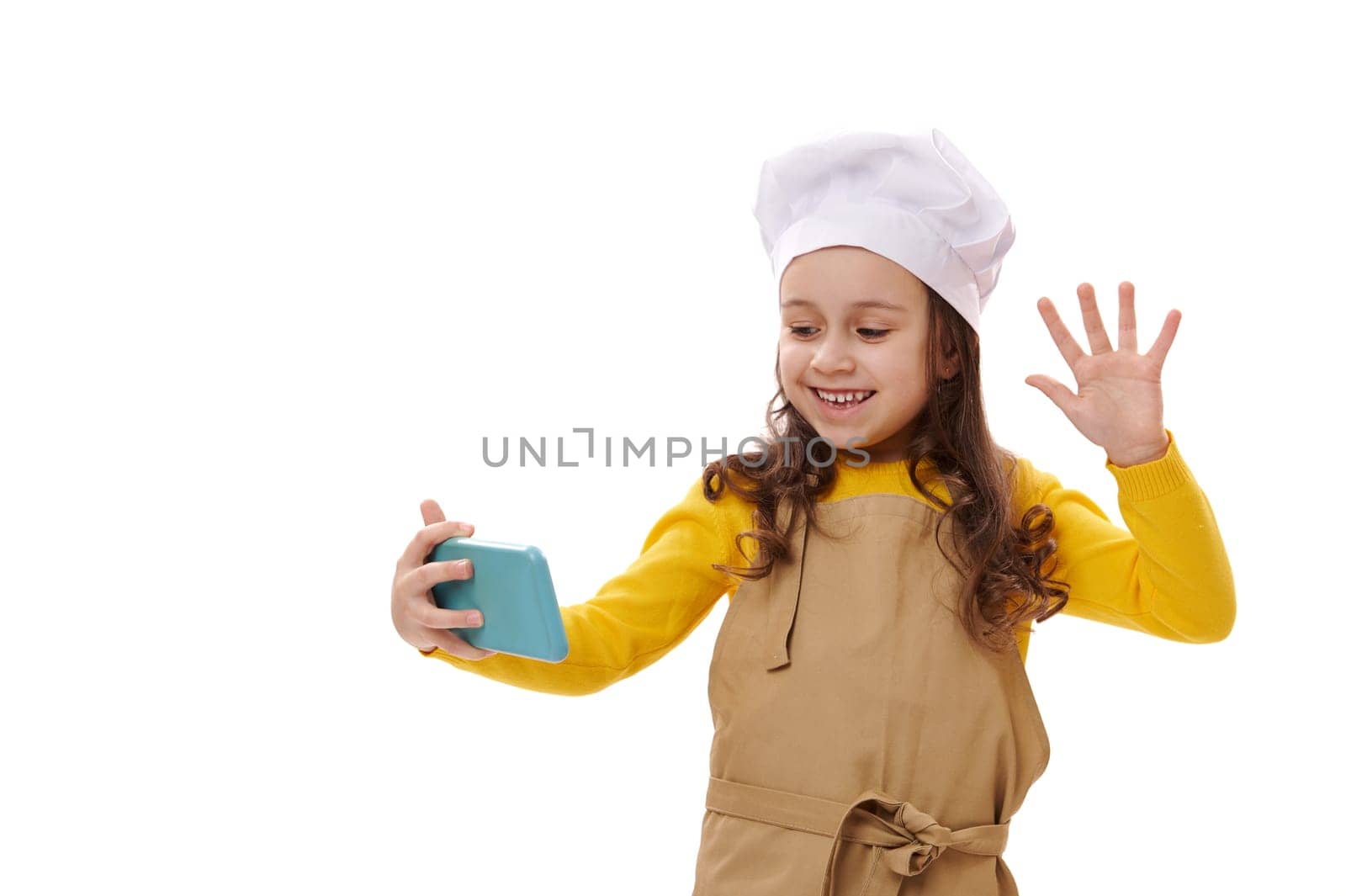 Smiling positive 5-6 years child girl in white chef's hat and beige kitchen apron waving with her hand while communicating online during video conference, isolated over white background. Copy ad space