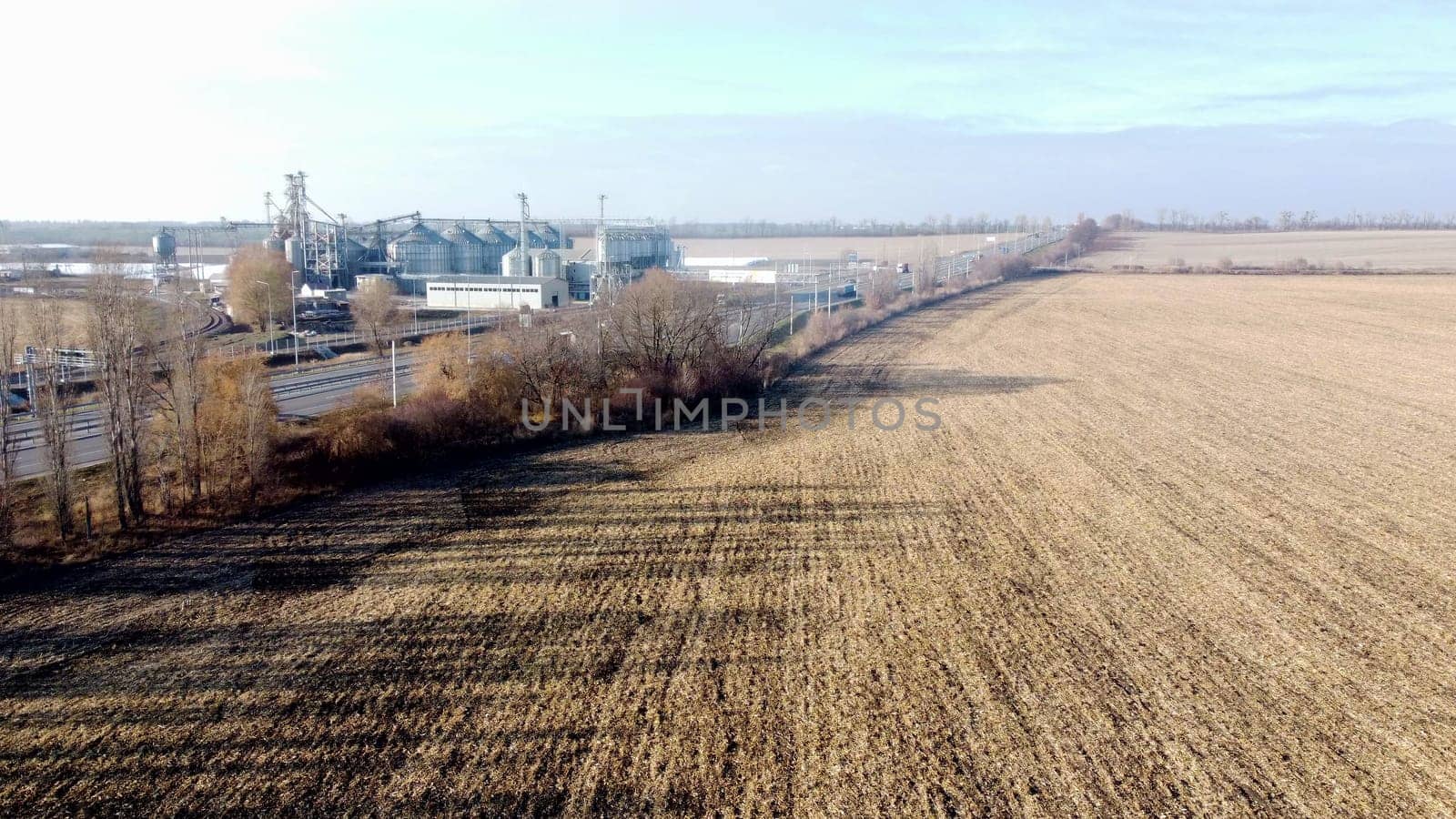 Agricultural industrial landscape. Plowed field after harvesting dry wheat, an elevator, a highway with moving cars on an autumn sunny day. Yellow field with dug up earth, road, cars, grain elevator