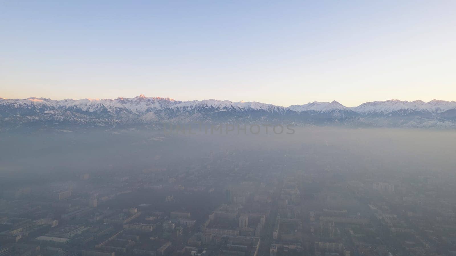 Epic gray smog is visible at sunset over the city. A bird's-eye view from a drone of houses, roads, cars and parks. White clouds and snowy mountains are illuminated by orange rays of the sun. Almaty