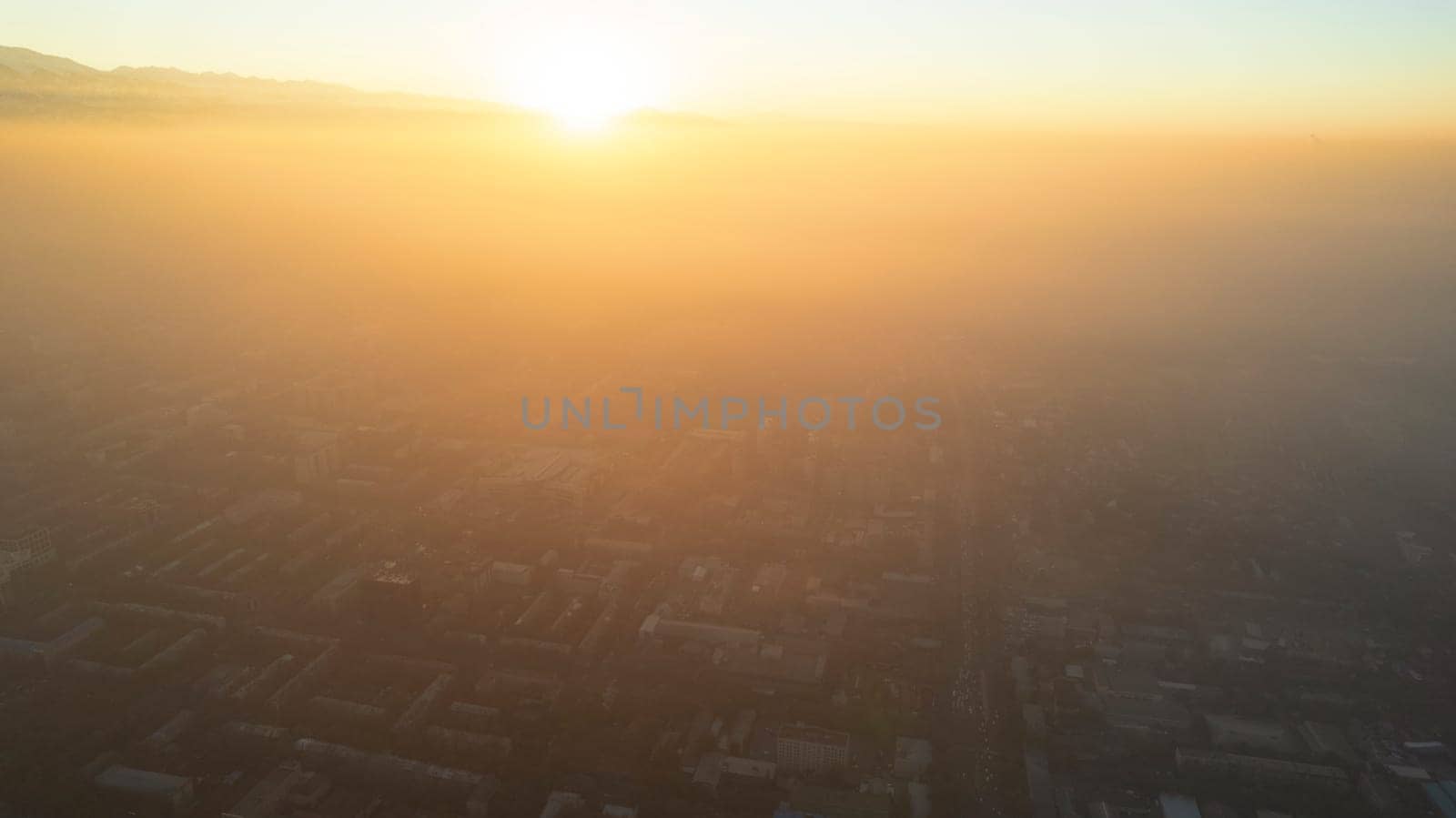 Epic gray smog is visible at sunset over the city. A bird's-eye view from a drone of houses, roads, cars and parks. White clouds and snowy mountains are illuminated by orange rays of the sun. Almaty