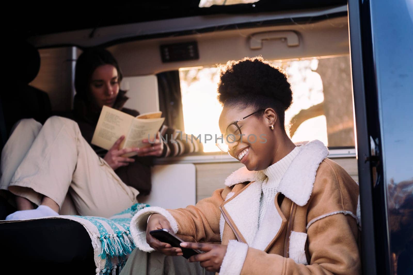 smiling african woman chilling checking phone at sunset during a road trip in camper van with a friend, concept of adventure travel with friend and technology of communication