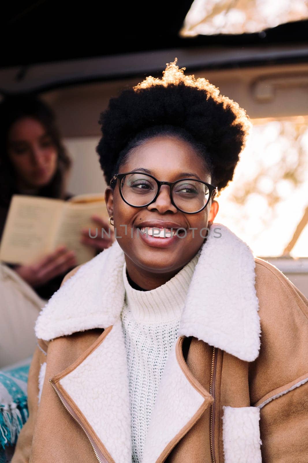 vertical portrait of a young black woman smiling at sunset sitting in a camper van with a friend reading in the background, concept of travel adventure and female friendship