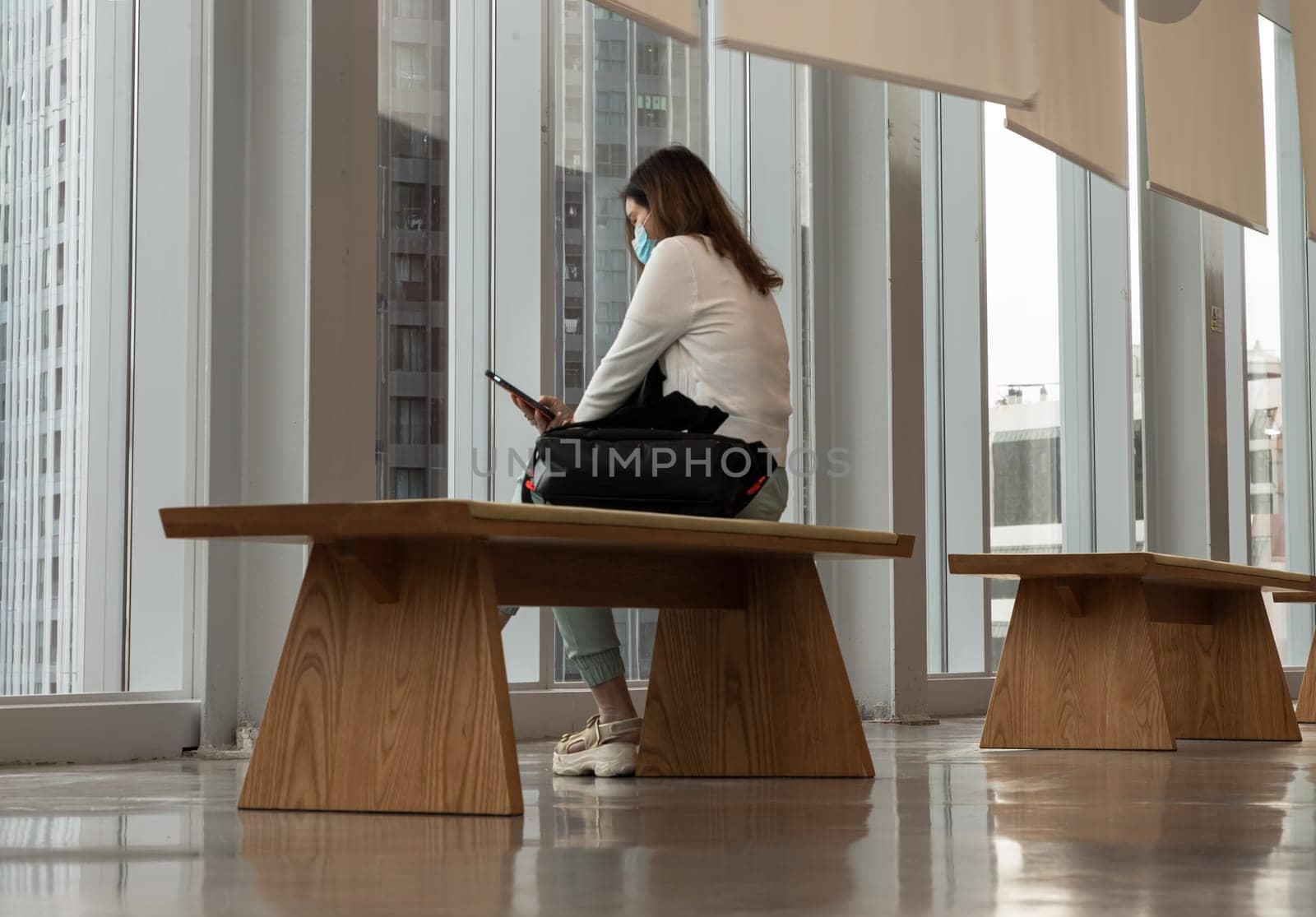 Bangkok,Thailand - Aug 13, 2022 - A calm young asia woman sitting on Modern wood bench wearing face mask protection for virus coronavirus covid 19 and using smart phone browse internet reading media news in empty hall room with glossy floor and cityscape. Side view, No focus, specifically.