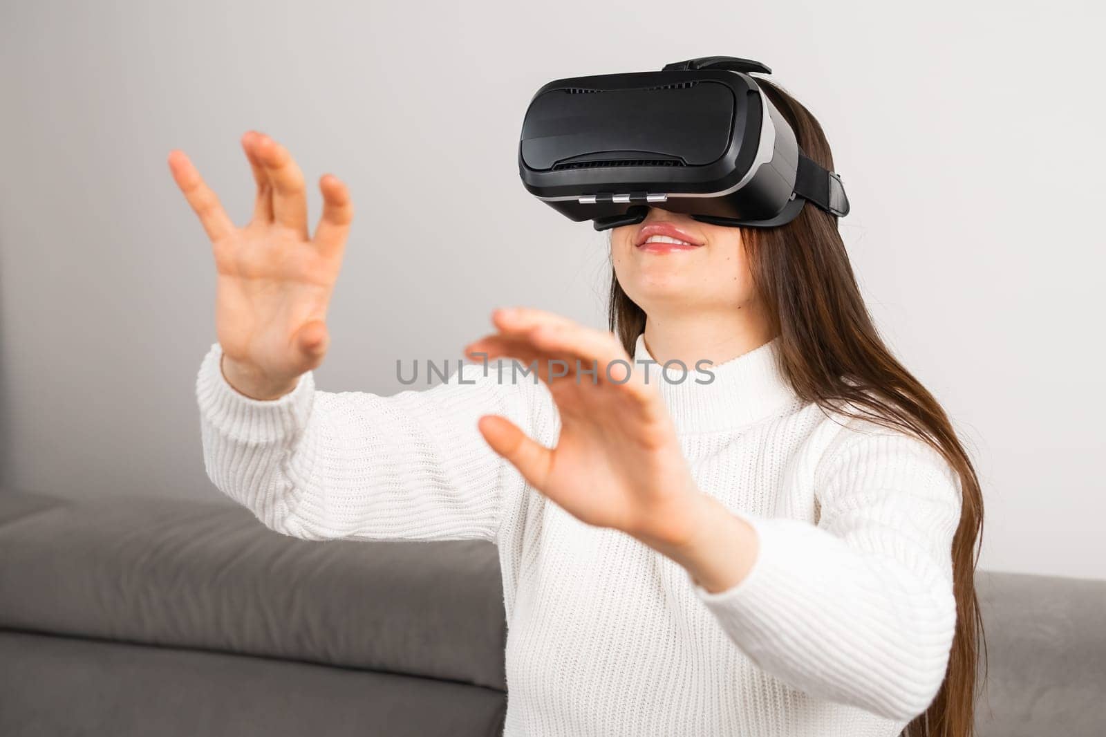 Curious woman swipes virtual interface with hands looking through VR glasses. Young housewife sits on stylish sofa enjoying visual effects at home