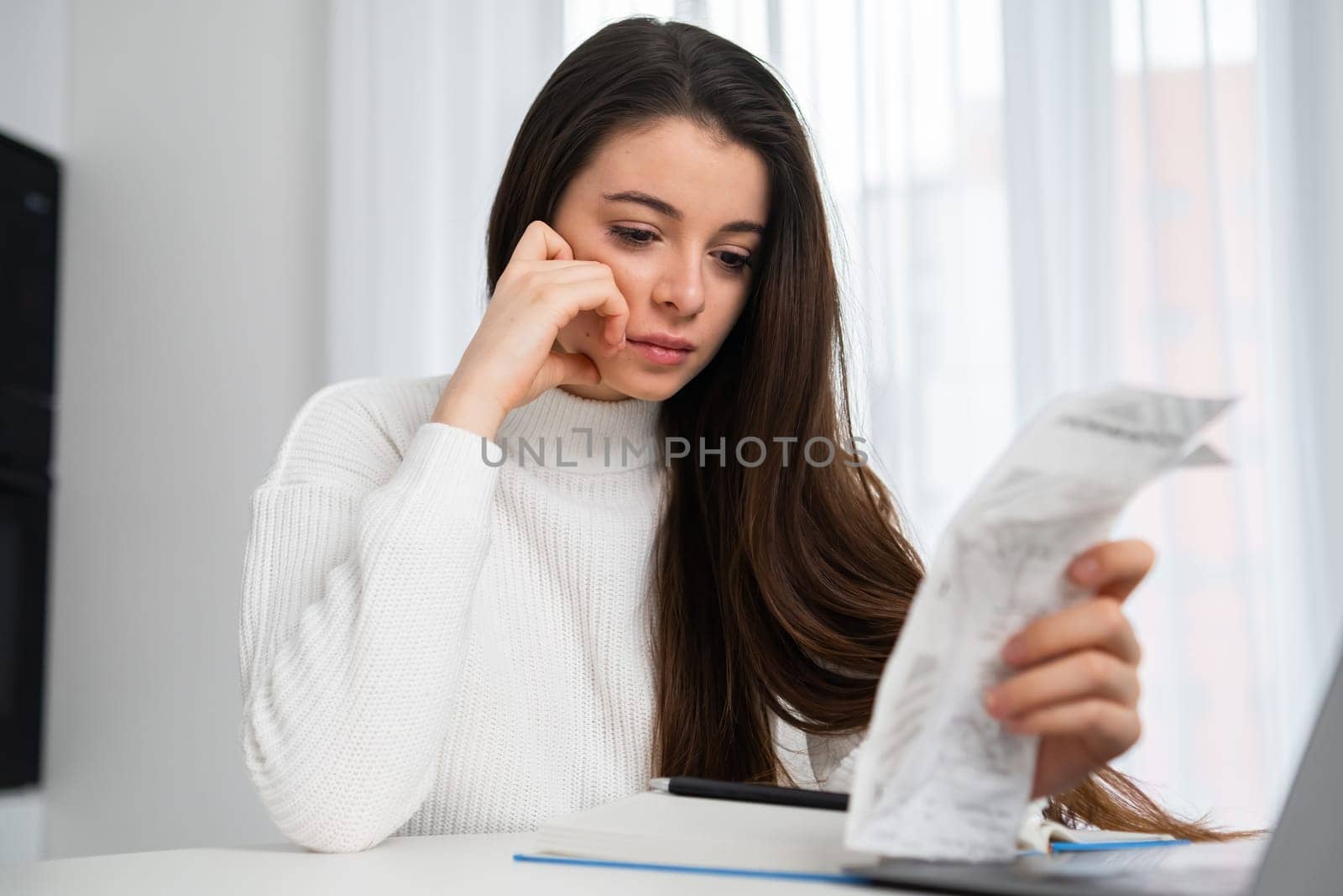 Sad woman regrets spending money on unnecessary purchases sitting at table near laptop in room. Brunette lady looks at long paper check from store