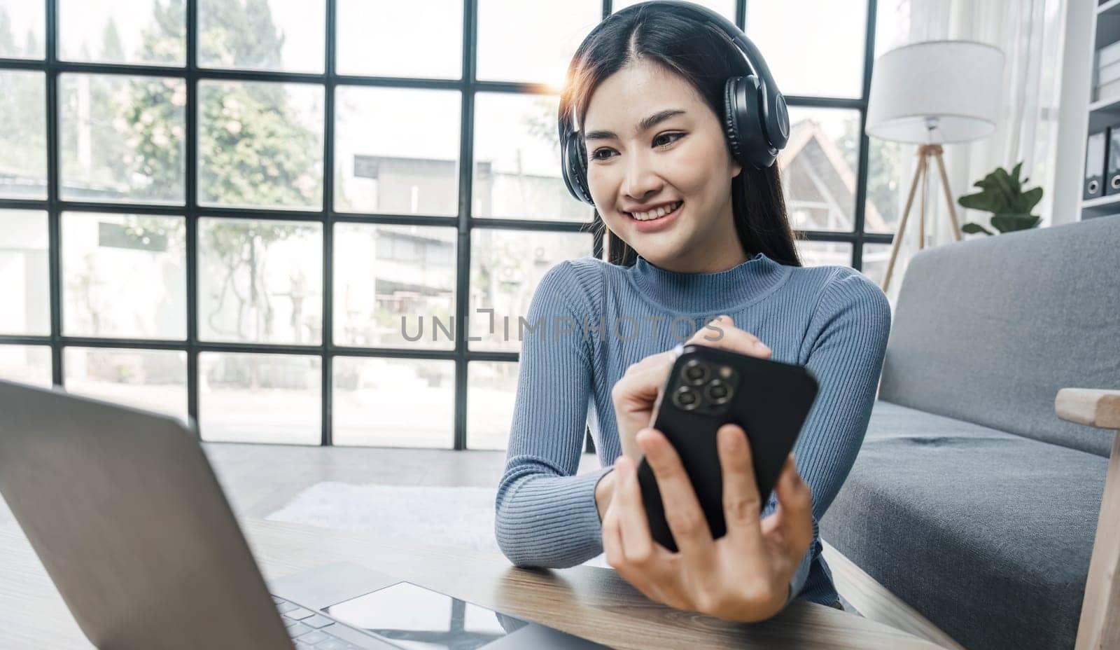 Smiling young asian woman using laptop web camera while wearing headphones and sitting on the rug beside to the sofa at homes..