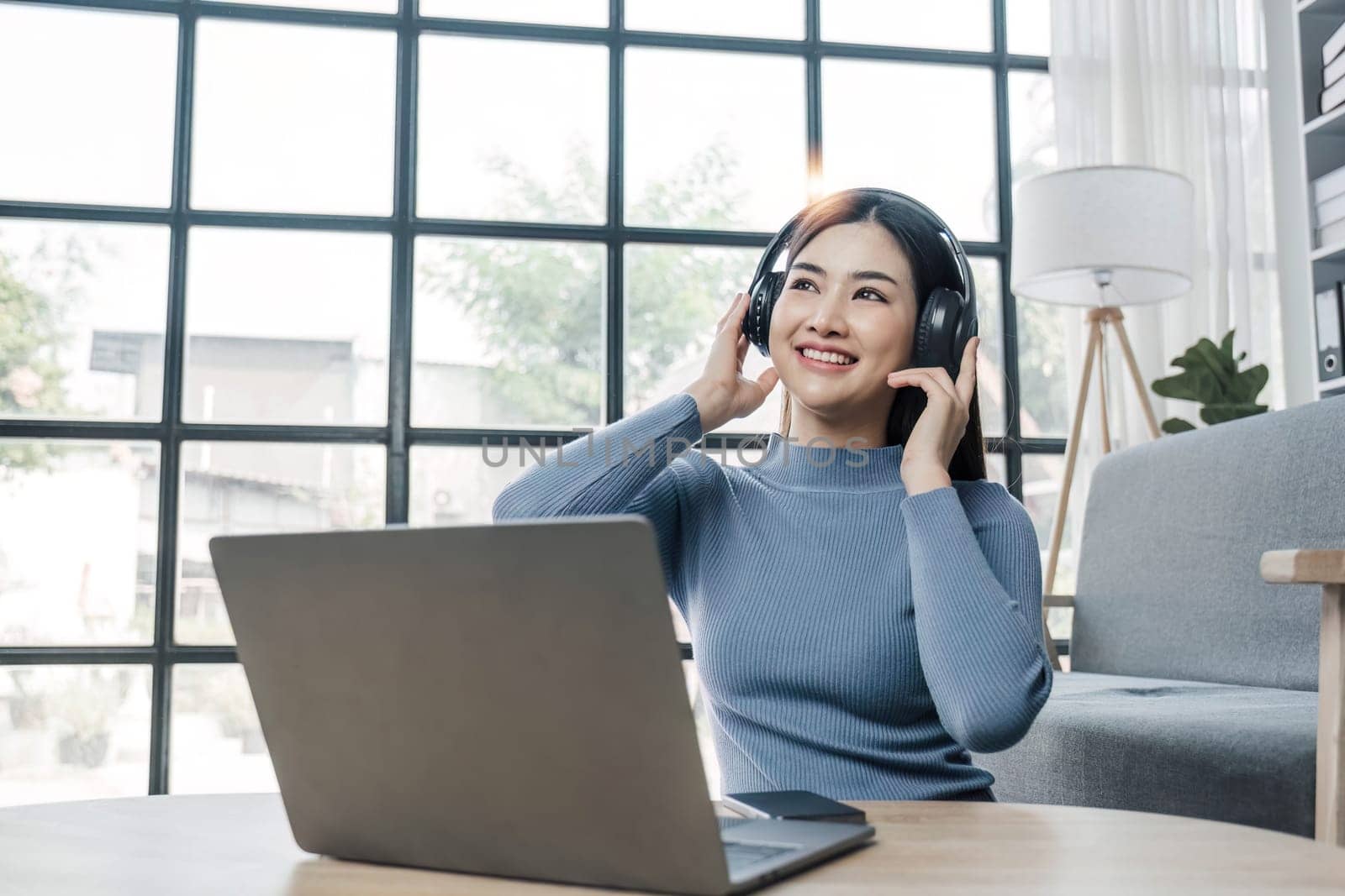 Smiling young asian woman using laptop web camera while wearing headphones and sitting on the rug beside to the sofa at homes..