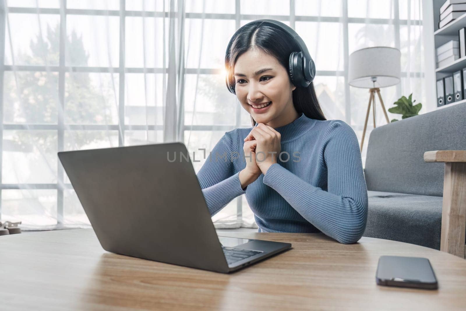 Smiling young asian woman using laptop web camera while wearing headphones and sitting on the rug beside to the sofa at homes..