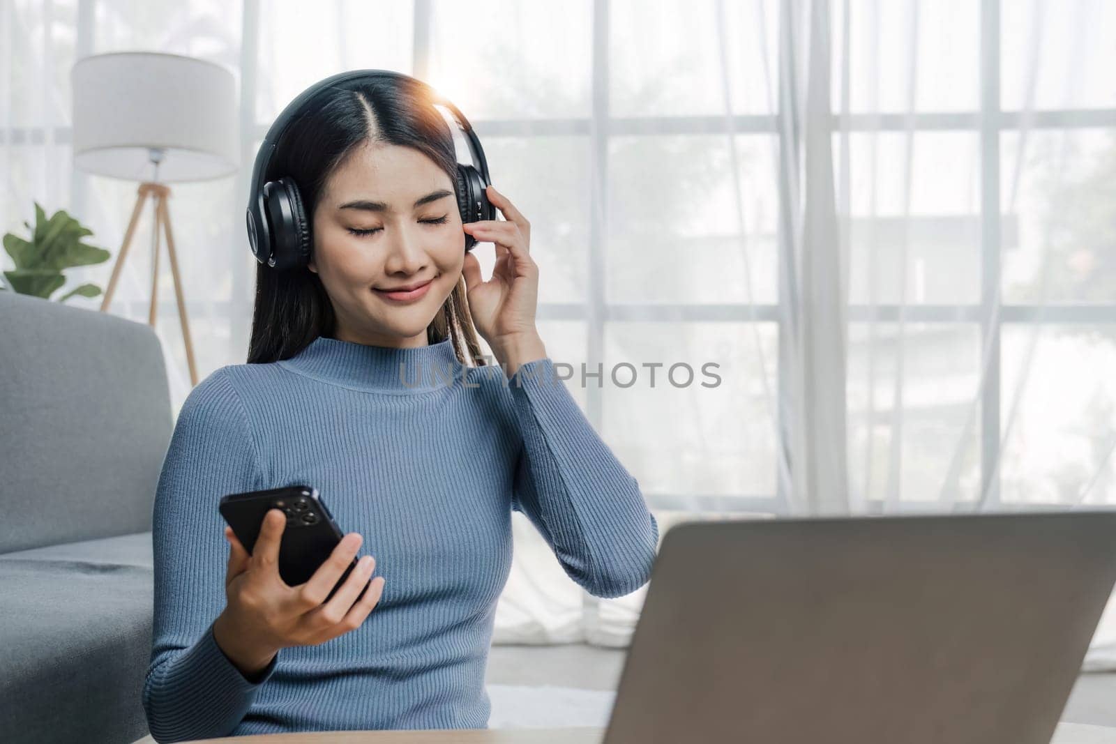 Smiling young asian woman using laptop web camera while wearing headphones and sitting on the rug beside to the sofa at homes..