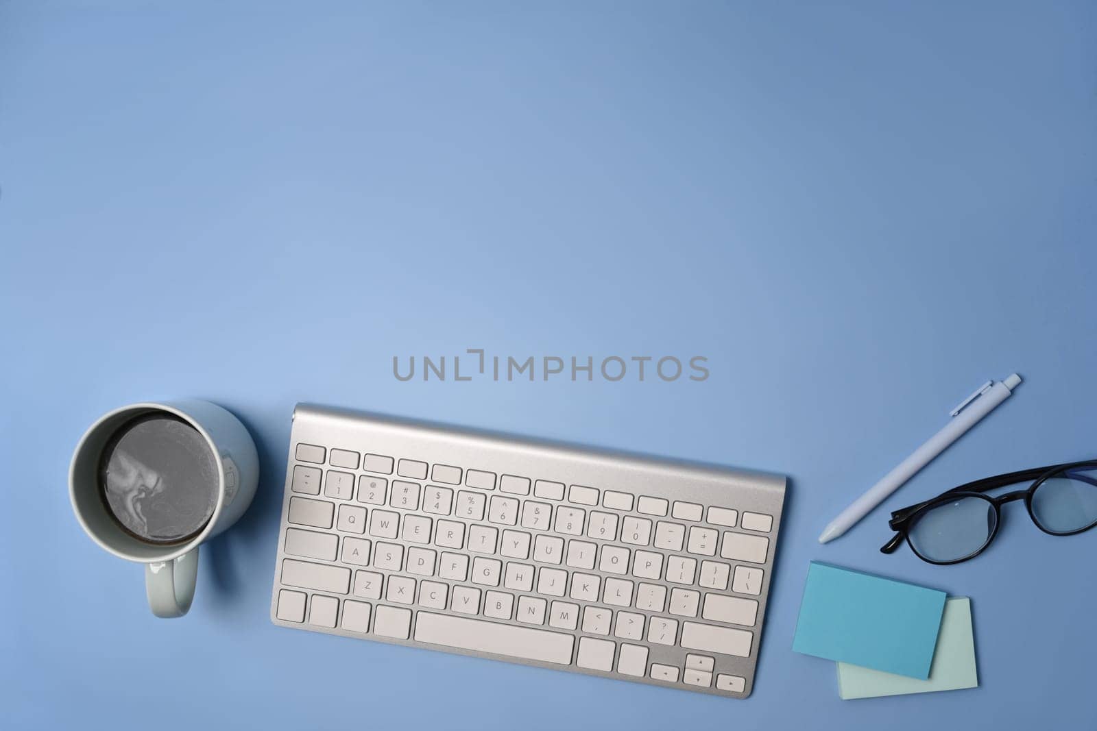 Top view wireless keyboard, coffee cup and glasses on blue background.