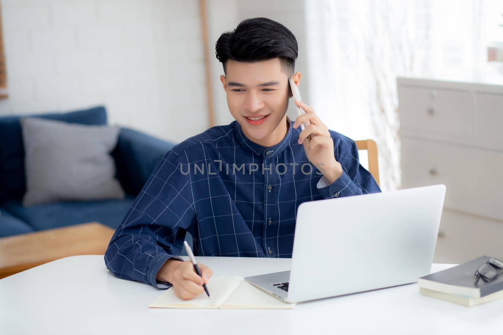 Young asian man talking phone and work from home with laptop computer, freelance male writing on notebook and speak on smartphone, stay home, domestic life, business and communication concept. by nnudoo