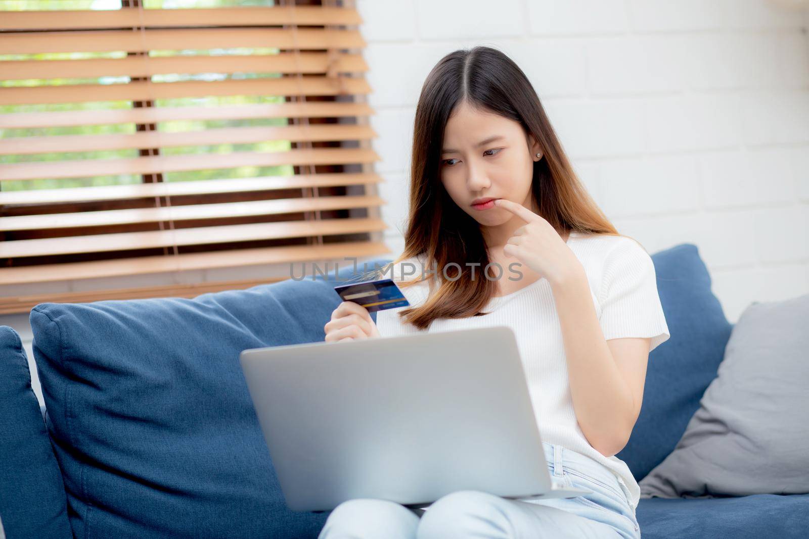 Young asian woman sitting thinking idea using credit card with laptop computer on couch, girl shopping online for buy and payment with notebook on sofa, finance and debit, lifestyle concept.