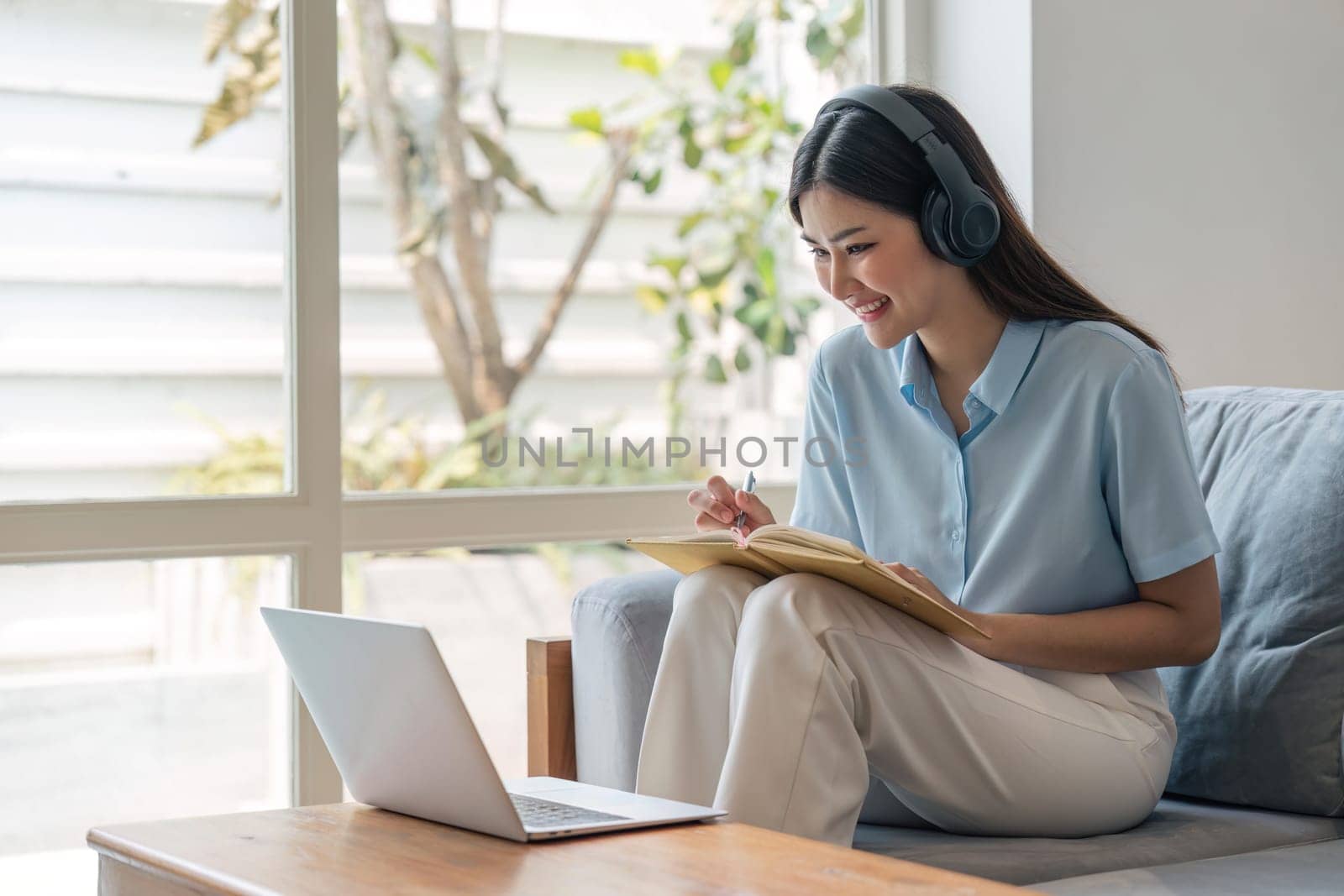 woman taking notes in notebook while using laptop at home in living room. Focused lady writing in notepad by nateemee