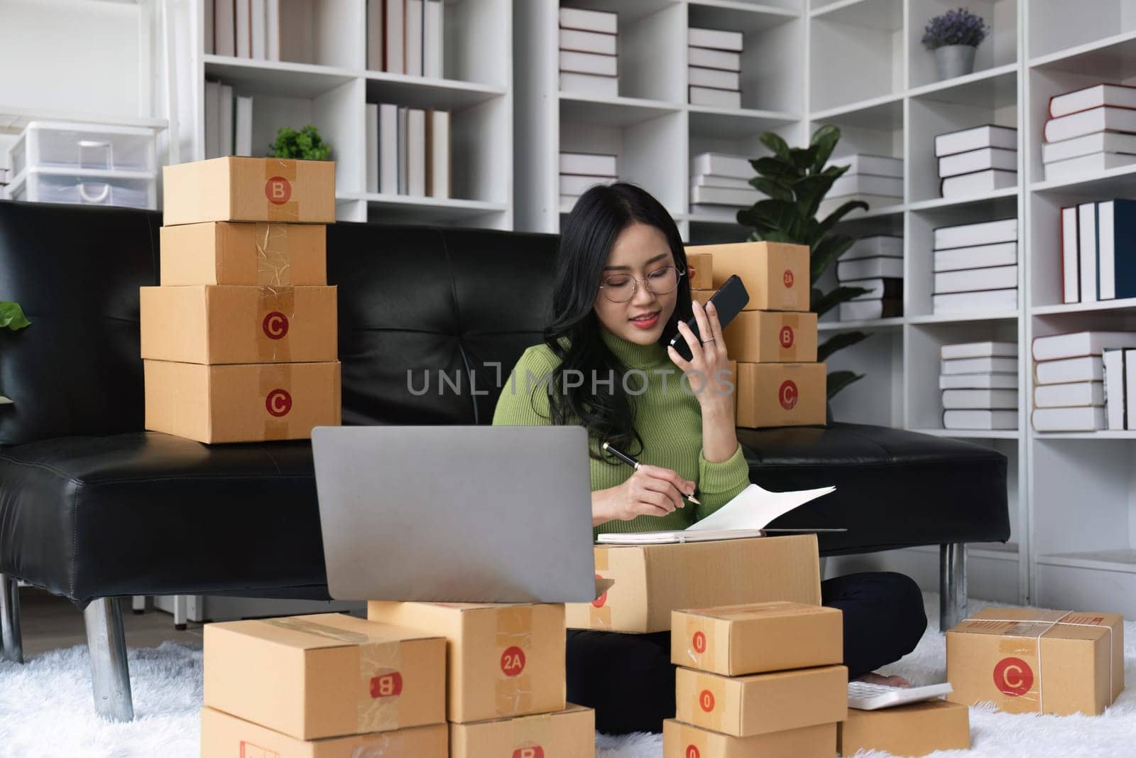 Happy Asian woman business owner working and selling online with computer laptop smile and talking with customer to get order in mobile phone. Small business concept.