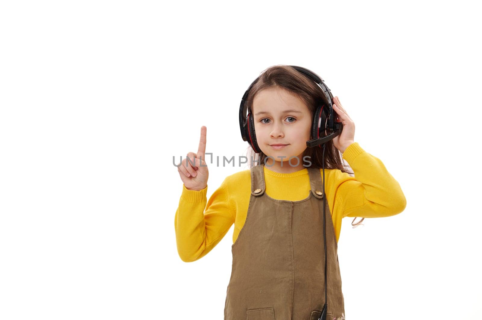 Beautiful little girl, first grader wearing headphones for online education or distance learning, pointing her index finger on a copy space, smiling looking at camera, isolated on white background.