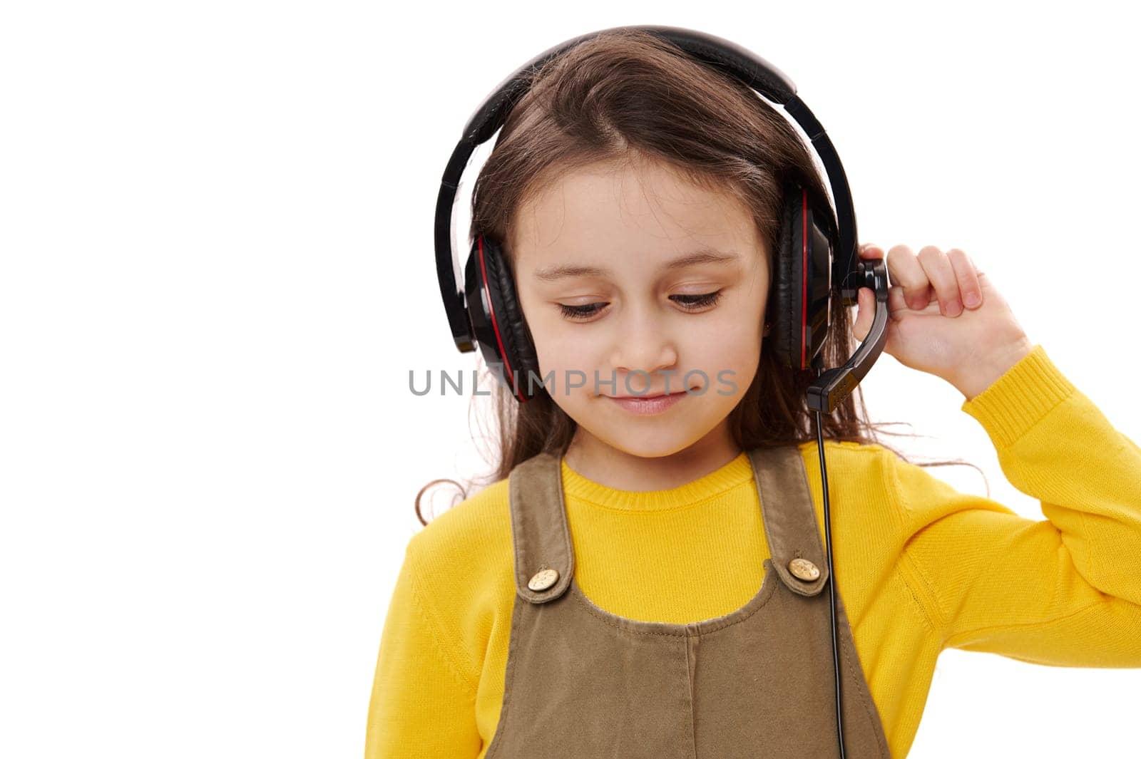 Mischievous little kid girl, primary school student, first grader with headphones, smiling over white isolated background. Smart pupil enjoys online lesson on audio headset. Distance education concept