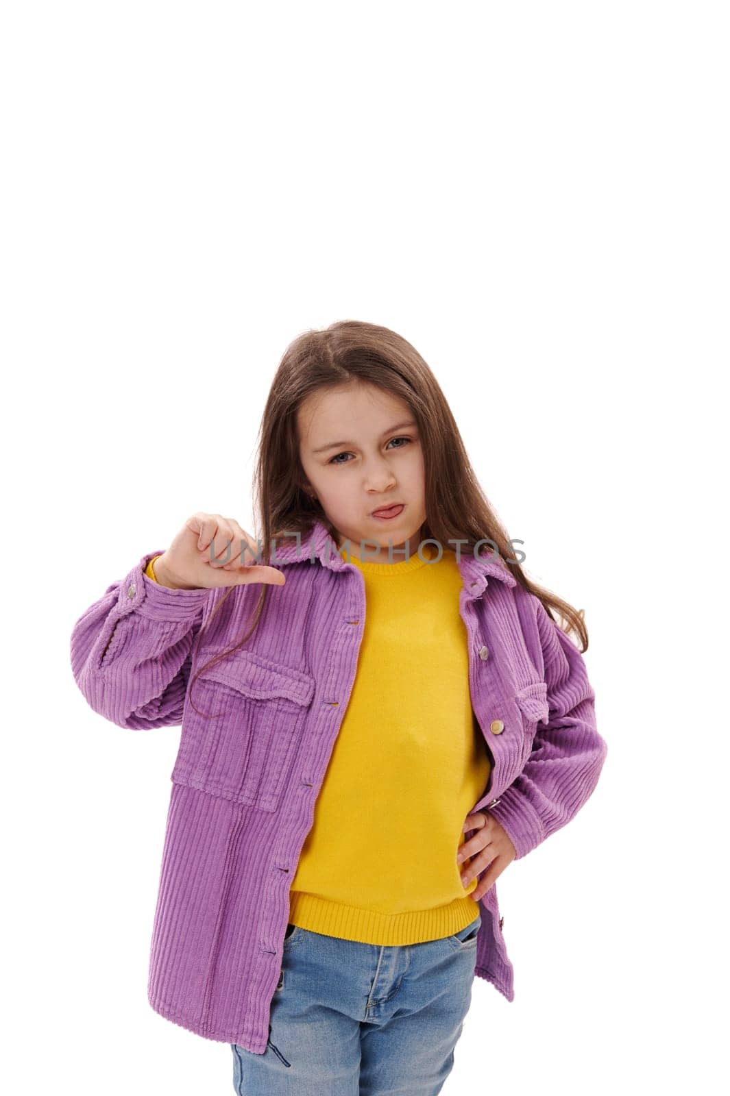 Mischievous 5-6 years little child girl gestures with thumbs down, makes faces, grimaces, shows her tongue looking at camera, expressing misunderstanding and disagreement, isolated on white background