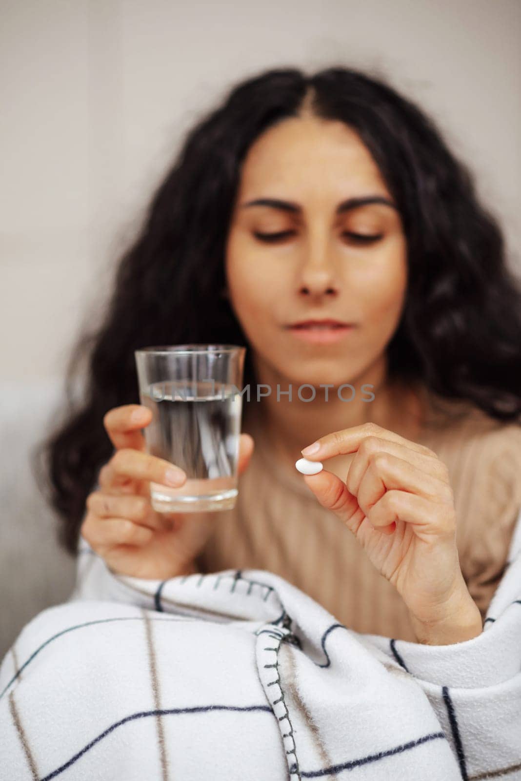 Beautiful young woman holding a pill and water. In the foreground a glass of water and a pill between the fingers. In the background is an Arab woman looking at a pill. The girl thought about taking medicine. Woman about to take medication. A young girl Got sick with the flu or a virus or possibly any form of allergy. A pill, a glass of water and a warm blanket is all she needs right now.