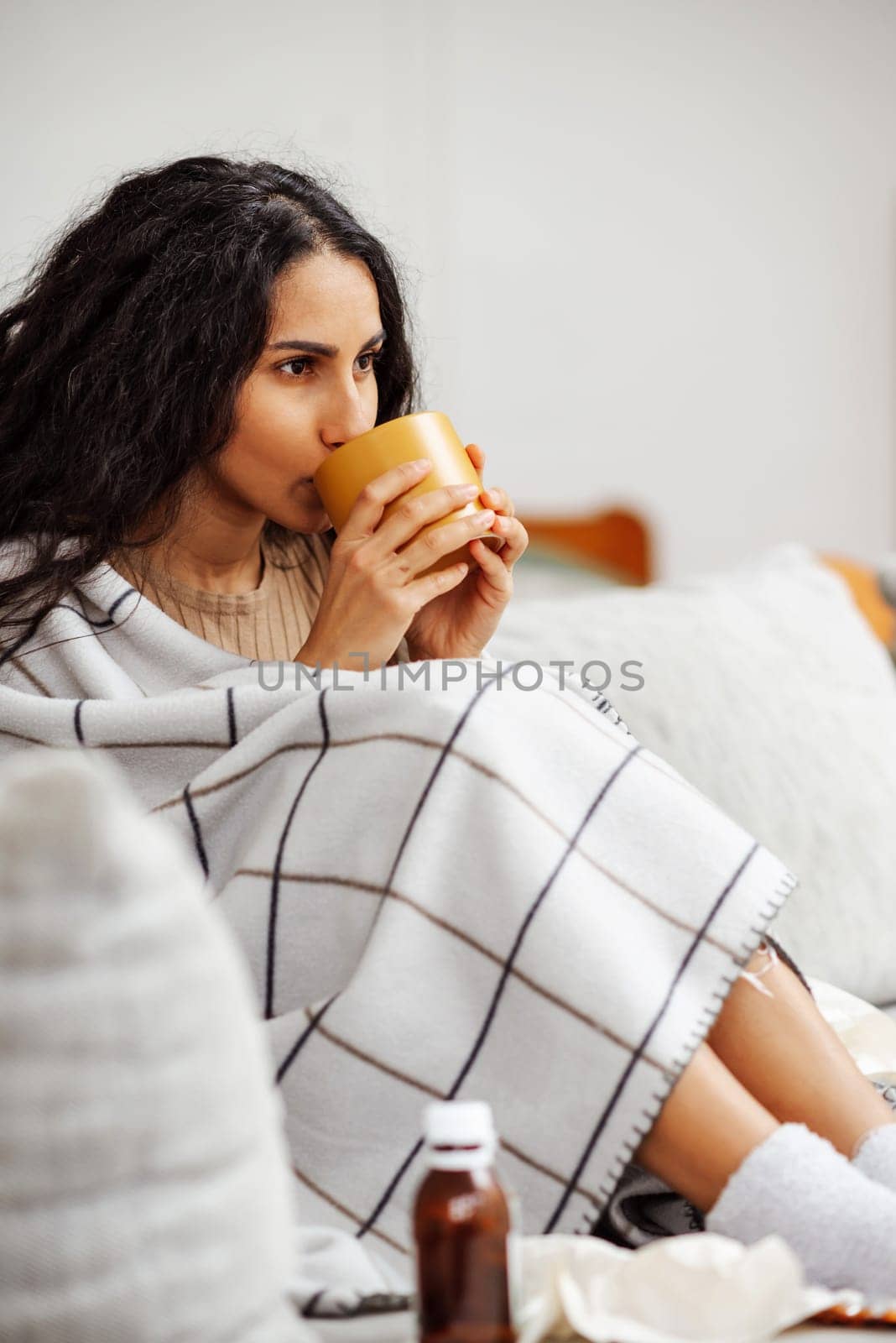 Beautiful young Arab girl drinks a cup of hot drink. Woman is wrapped in a blanket and sits on a gray sofa near her there are a lot of medicines. Recovery at home is always comfortable and safe.