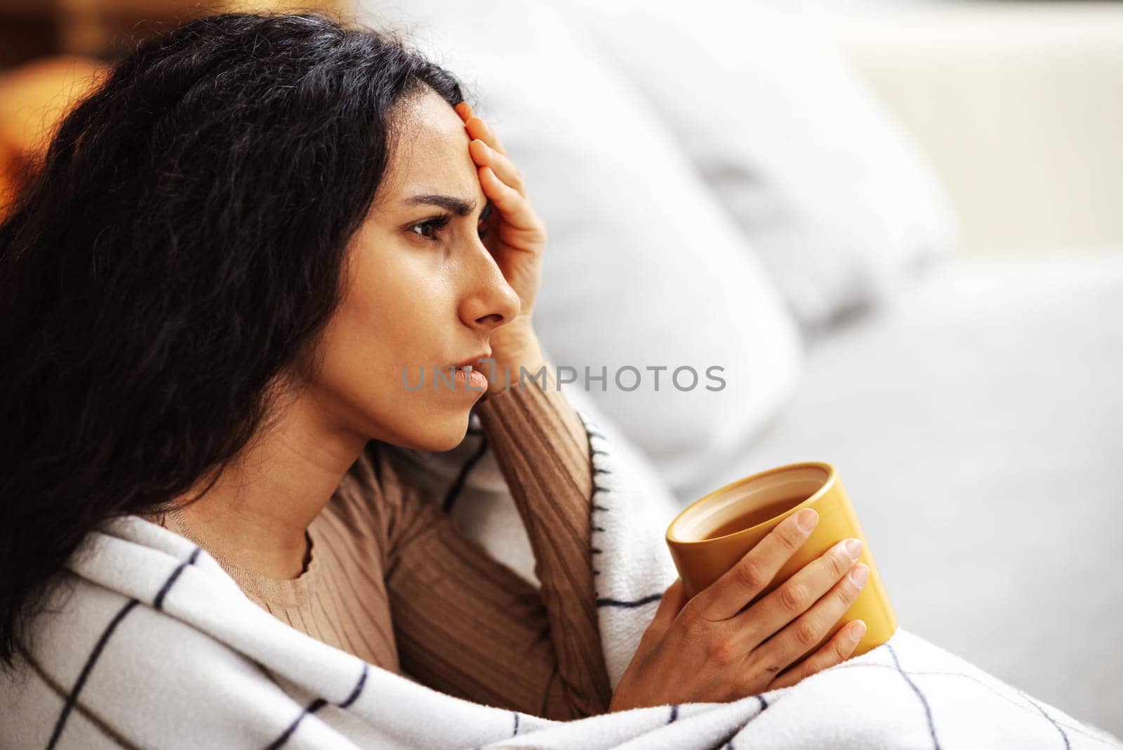 Young beautiful woman of Arab is sitting on a gray sofa wrapped in a blanket in one hand holding a cup with the other hand holding her head. Woman coping with stress and depression problems.