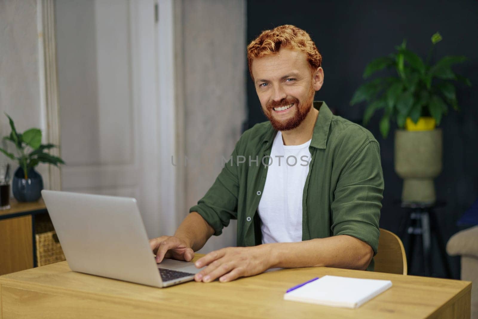Caucasian man with foxy hair sitting at desk in a cozy and comfortable home interior. He is using laptop to work from home, emphasizing the concept of remote work and the benefits of flexibility and independence. High quality photo