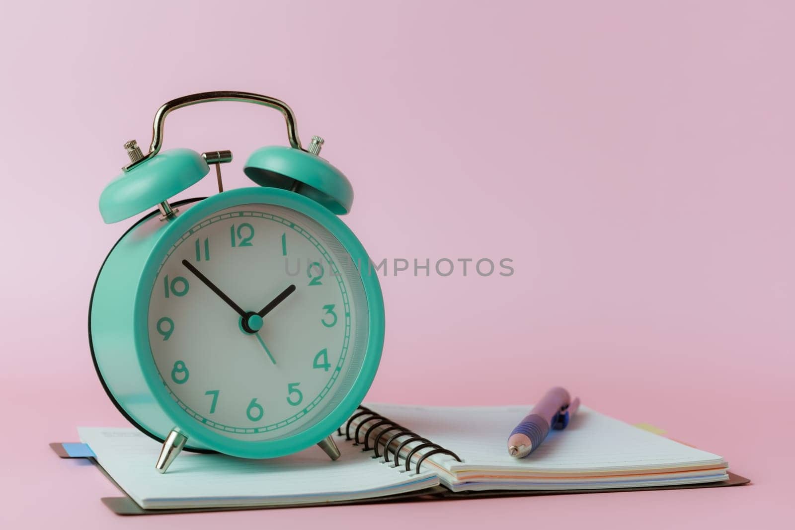 A bell alarm clock with a notebook and a pen on pink background for the concept of time management for work and study.