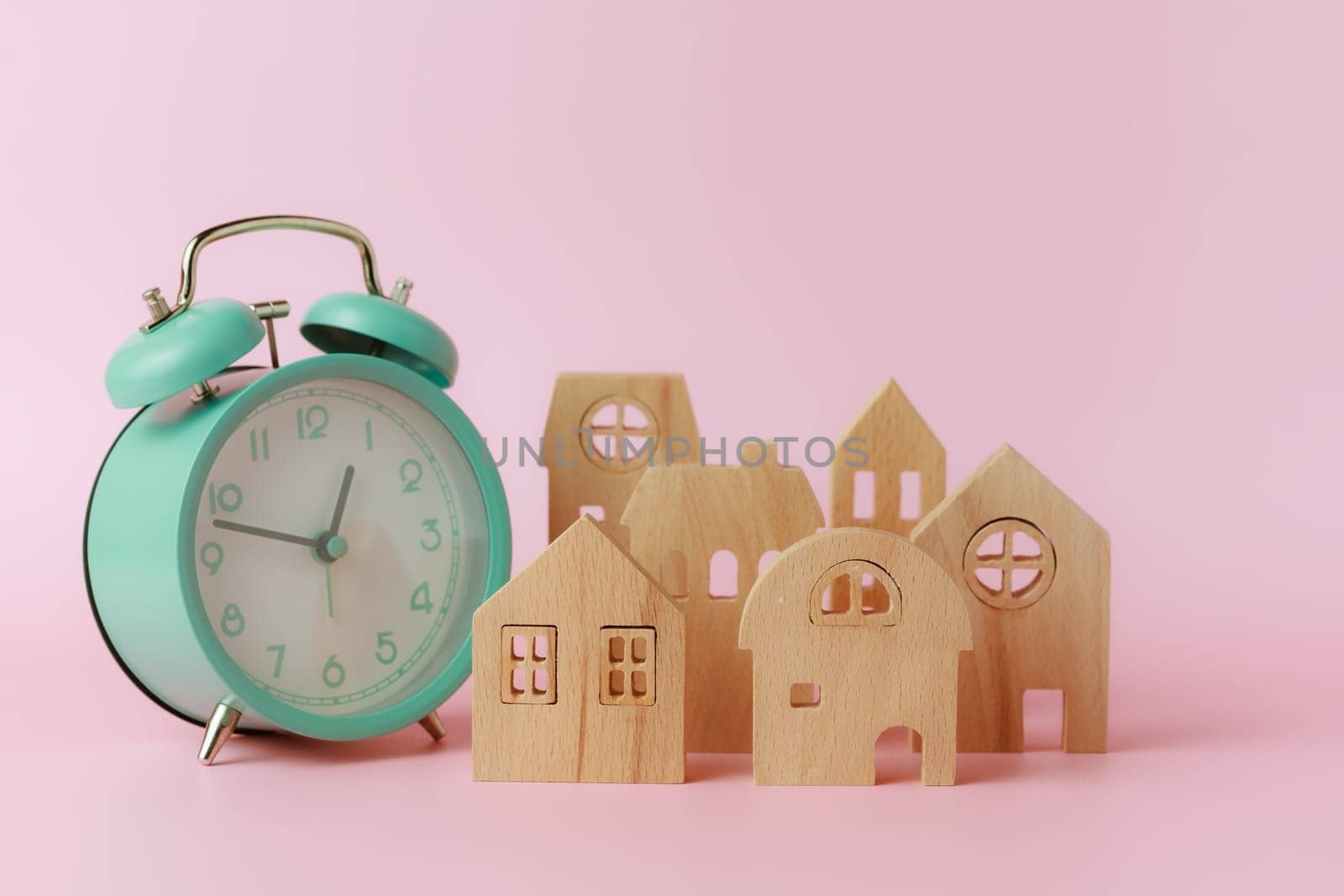 A bell alarm clock with wooden house model on pink background for the concept of time management, housing and property.