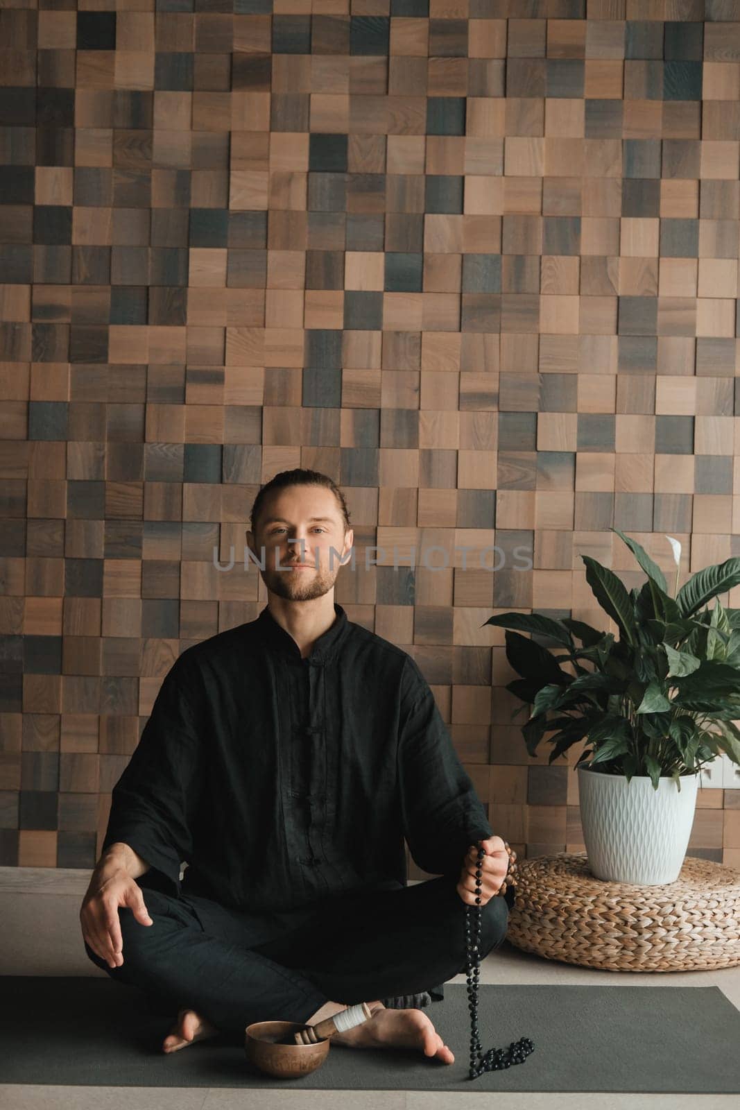 Portrait of a young man in a black kimano sitting in a lotus position on a gym mat in the interior.