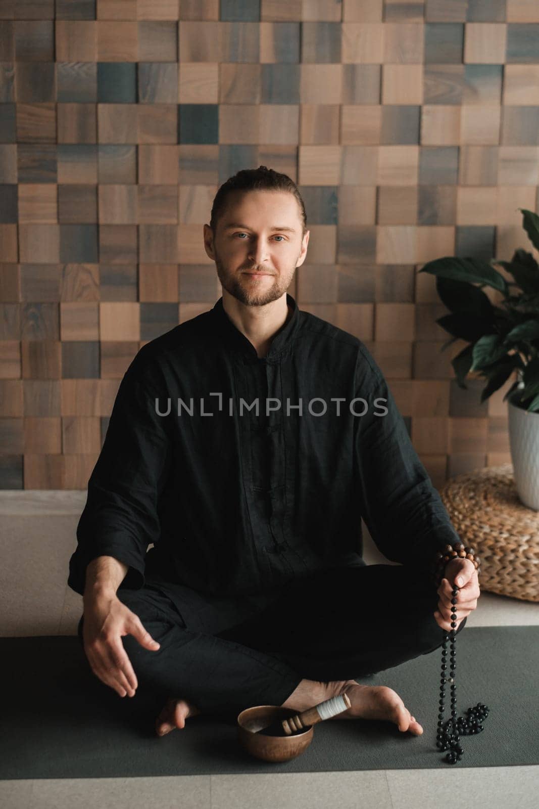 Portrait of a young man in a black kimano sitting in a lotus position on a gym mat in the interior by Lobachad